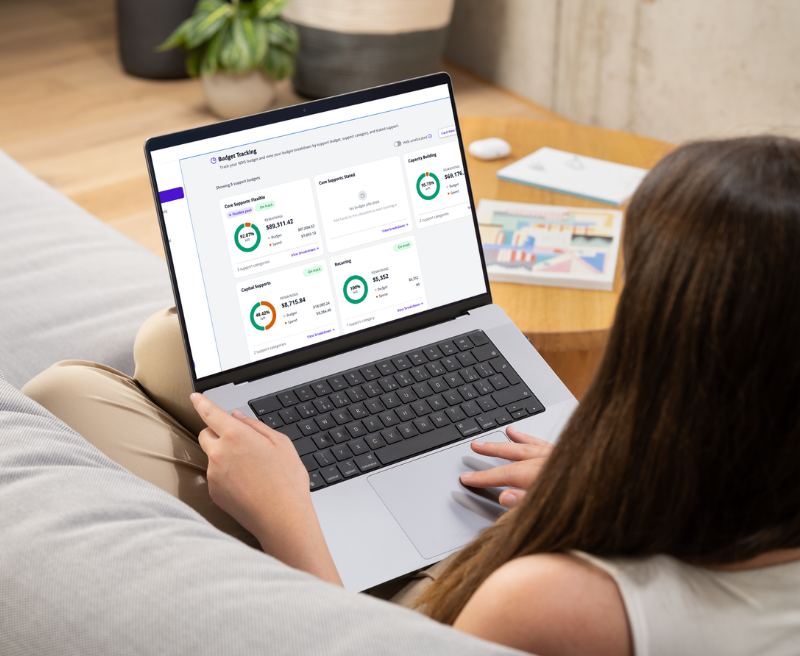 A woman on a couch with a laptop showing the Hai Helper Budget Tracking dashboard, with Core Supports, Capacity Building and Capital Supports categories broken down